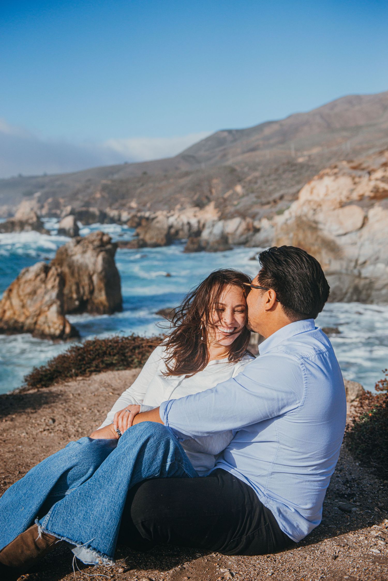 Couple embracing on rocky coastal cliffs overlooking dramatic Big Sur coastline with turquoise waves and green hills.