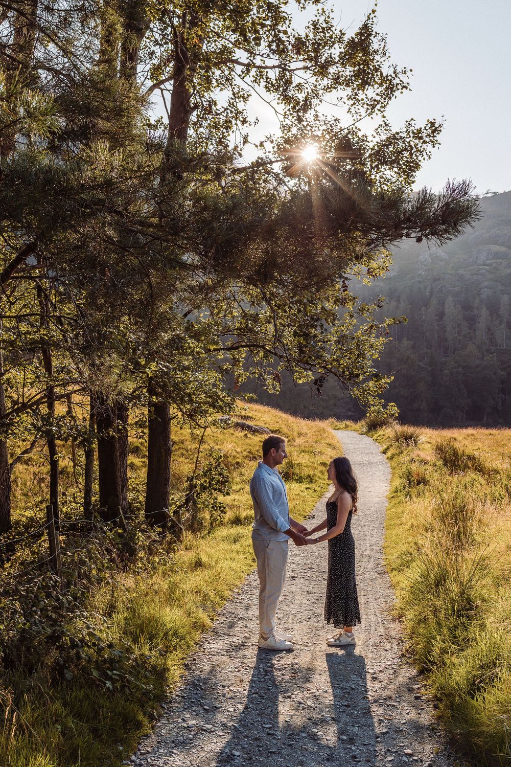 A couple stands together on a sunlit mountain trail surrounded by pine trees and golden grass at sunset.