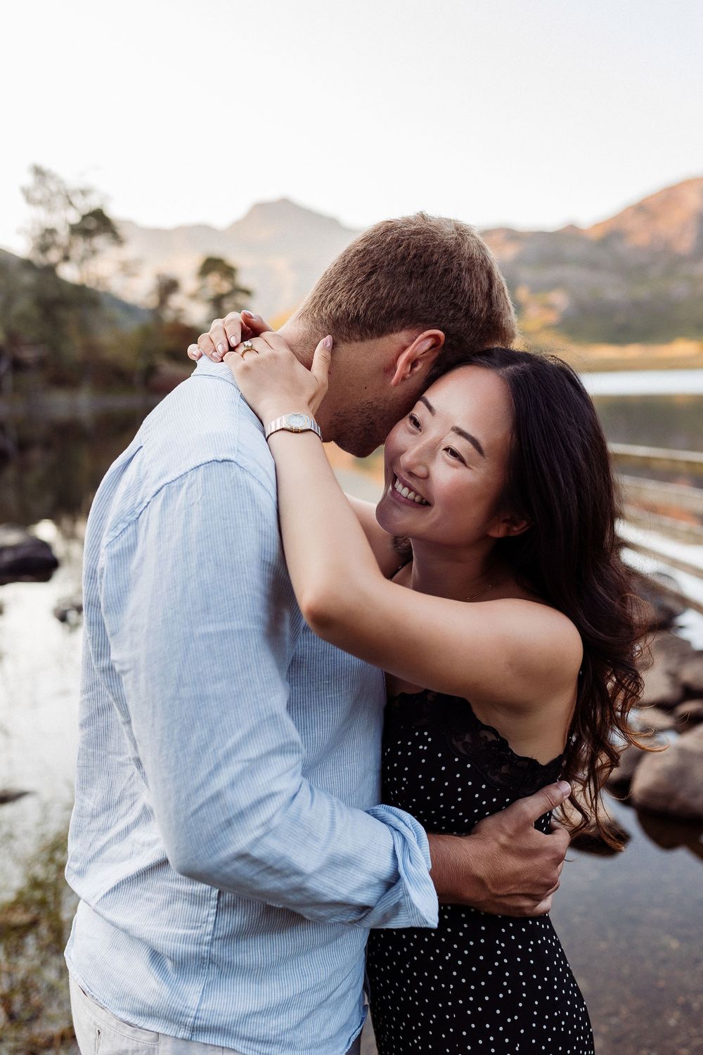 Couple shares joyful moments by a scenic lake with mountains in the background at sunset.