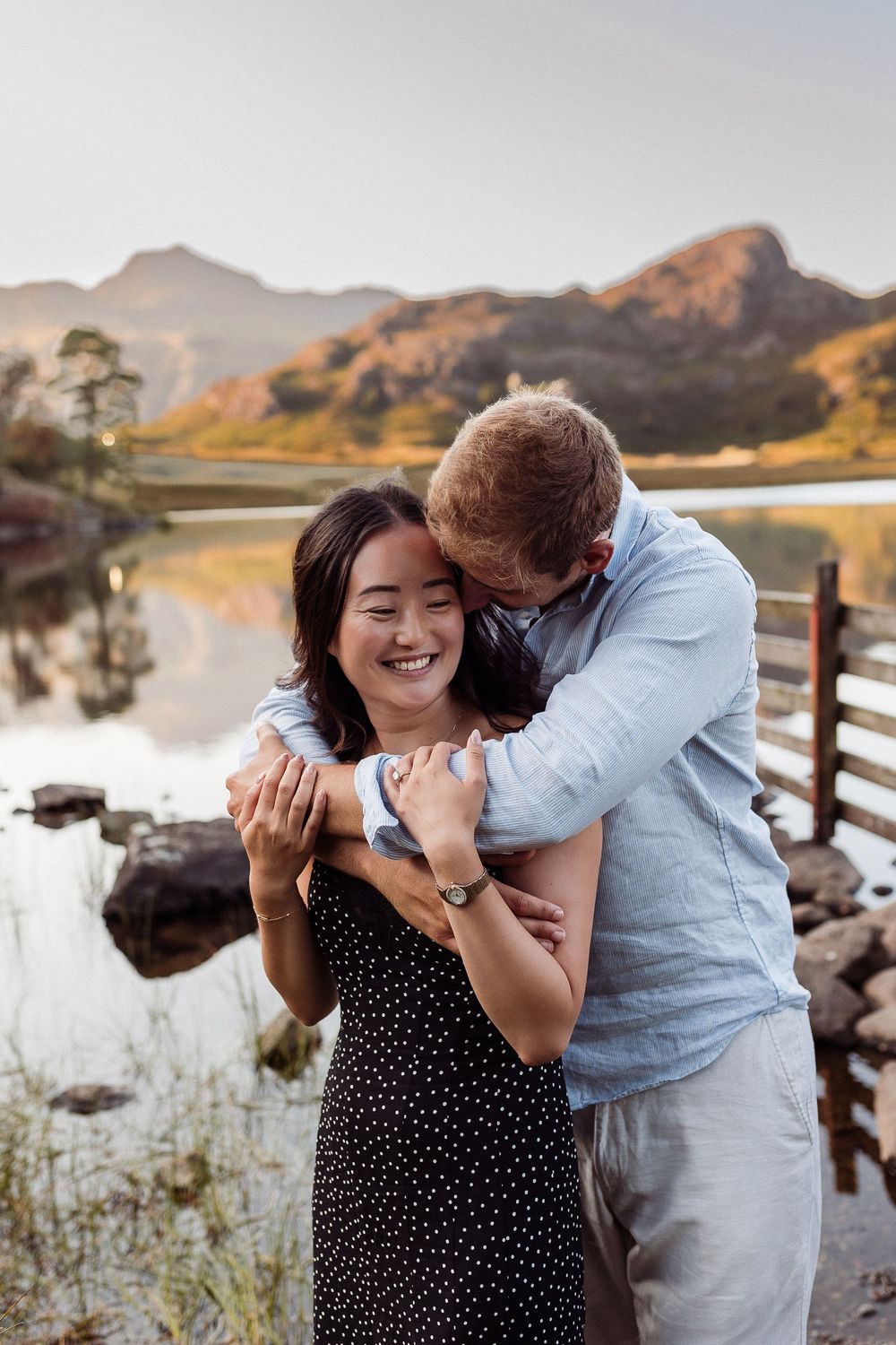 Romantic couple embraces and laughs together by a scenic mountain lake during sunset.