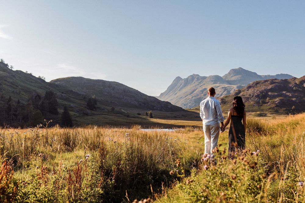 People hiking through golden grassland with scenic mountain views in the distance against a clear blue sky.