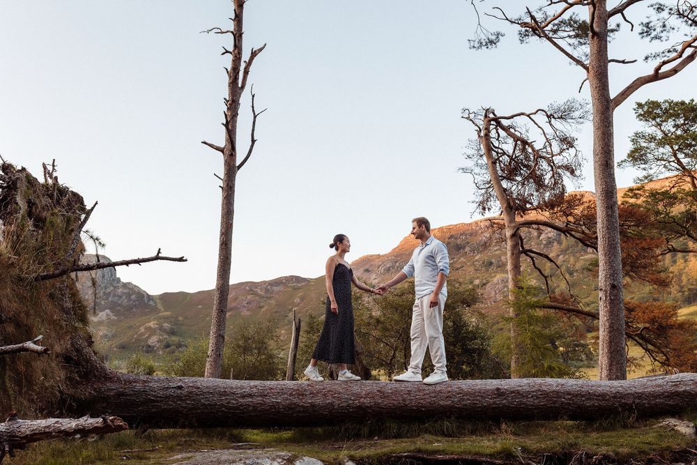 Two people carefully walk on a fallen tree trunk against a scenic mountainous backdrop at sunset.