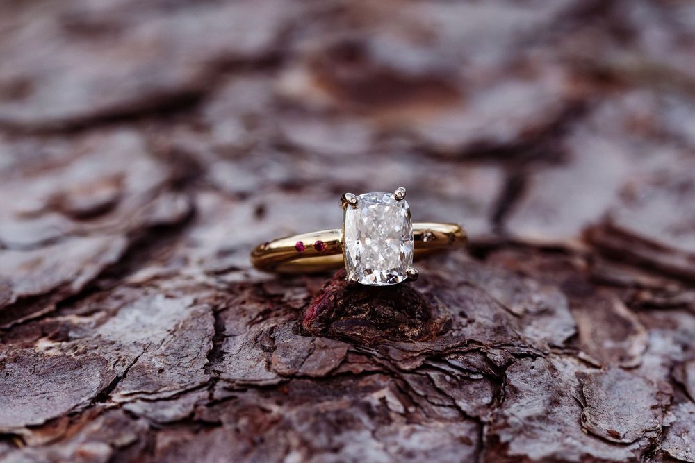 A radiant cut diamond engagement ring on gold band displayed on textured brown bark.