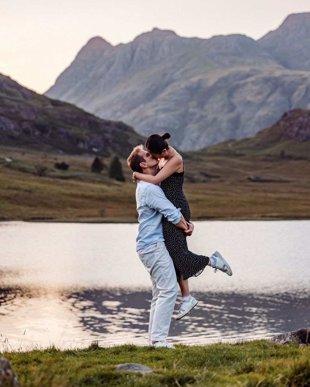 A couple embraces in a passionate lift against a backdrop of majestic mountains and a serene lake at dusk.