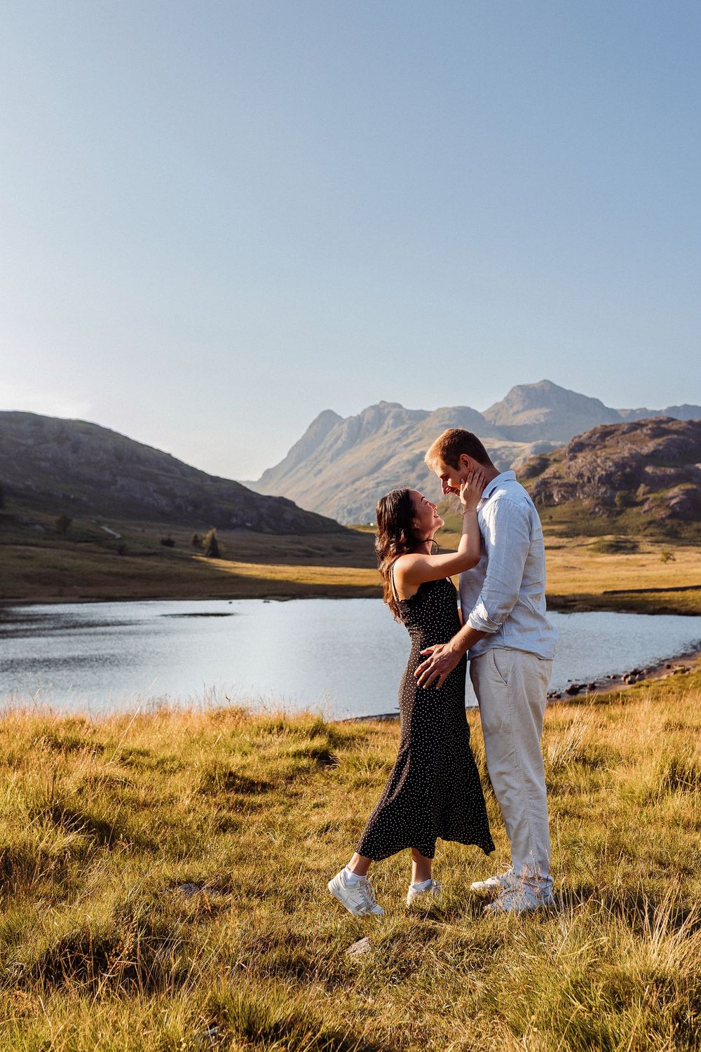 A couple embraces on a grassy hillside overlooking a serene mountain lake with peaks in the background.
