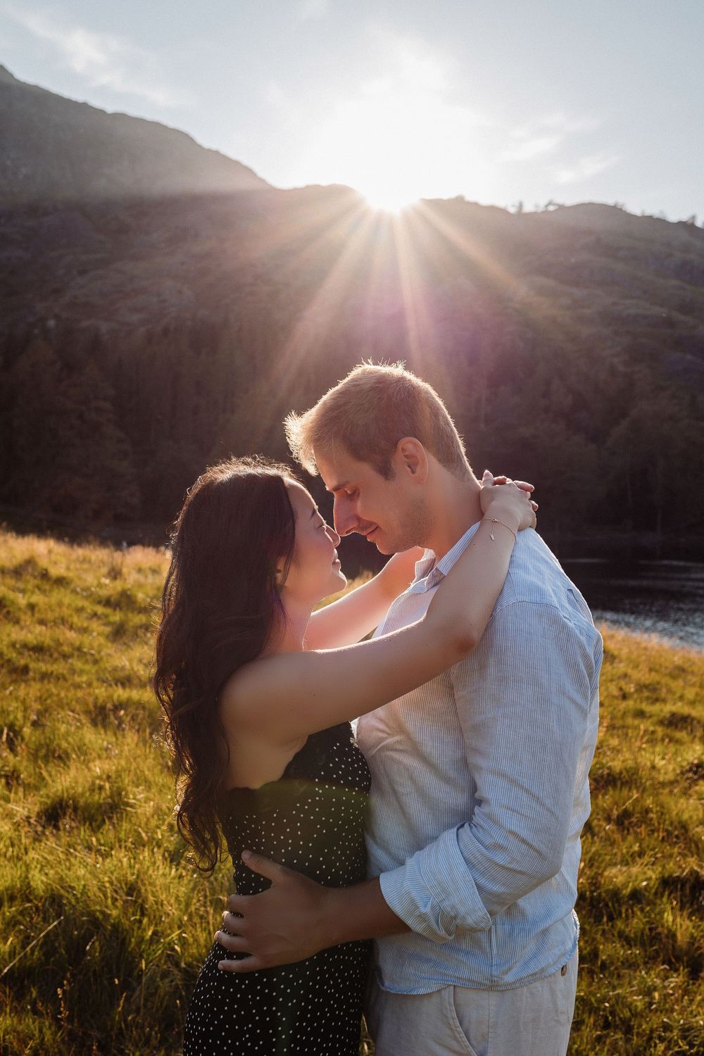 Romantic couple embraces in a sunlit meadow during golden hour with mountains in the background.