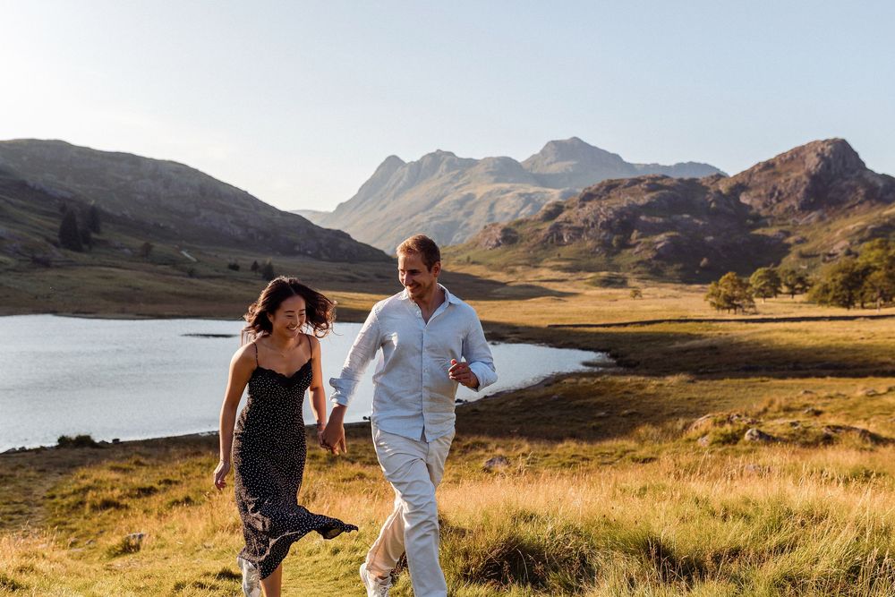 A couple strolls hand in hand on a golden hillside path beside a tranquil lake with mountains in the background.