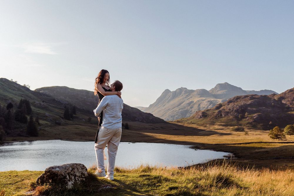 A silhouetted couple embraces on a hilltop overlooking a scenic mountain lake at sunset.