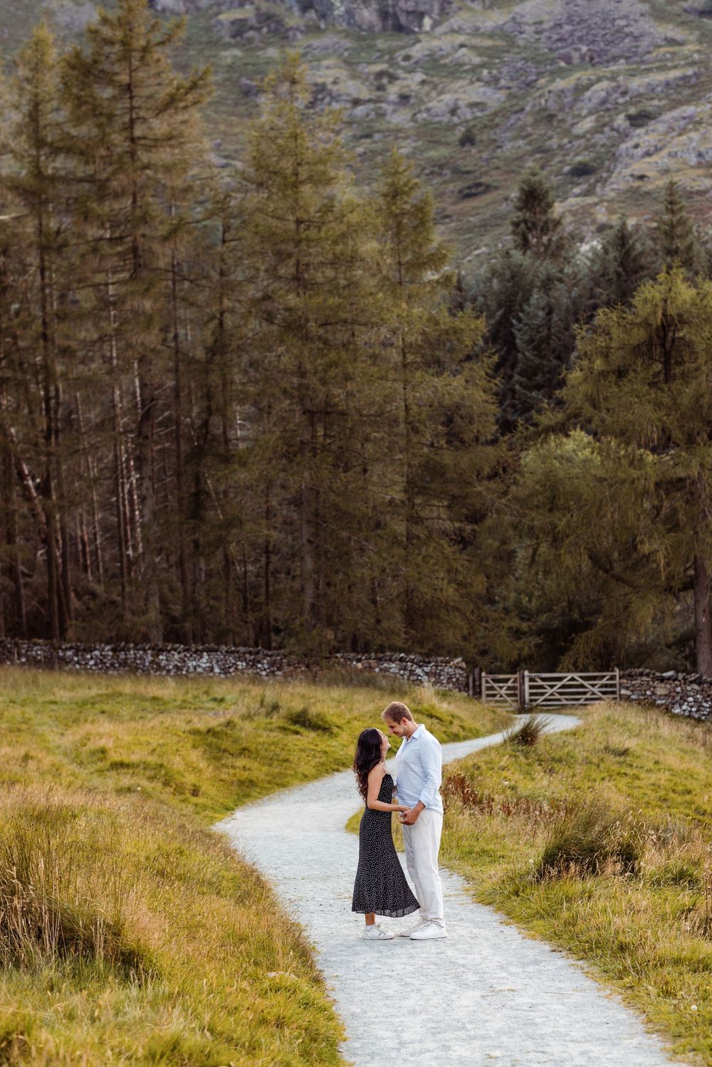 A couple walks along a winding mountain path through autumn trees and grassland in Lake District National Park.