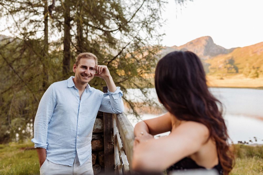 A couple takes photos of each other against a scenic mountain lake backdrop during golden hour.