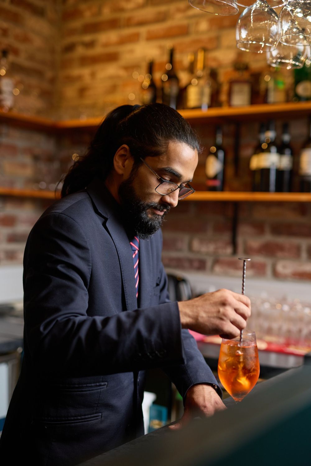 Person in formal attire examining a cocktail at a rustic brick-walled bar.
