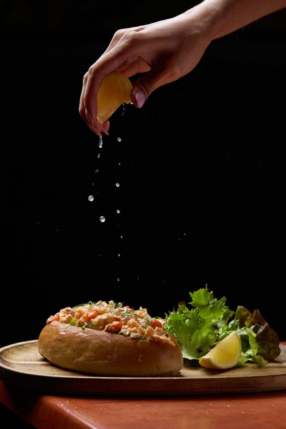 Hand squeezing citrus over an artfully plated sandwich with lettuce garnish on a brown plate.