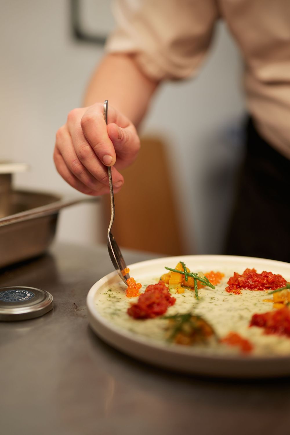 Close-up of a chef plating colorful garnishes and sauces on a white dish in a professional kitchen.