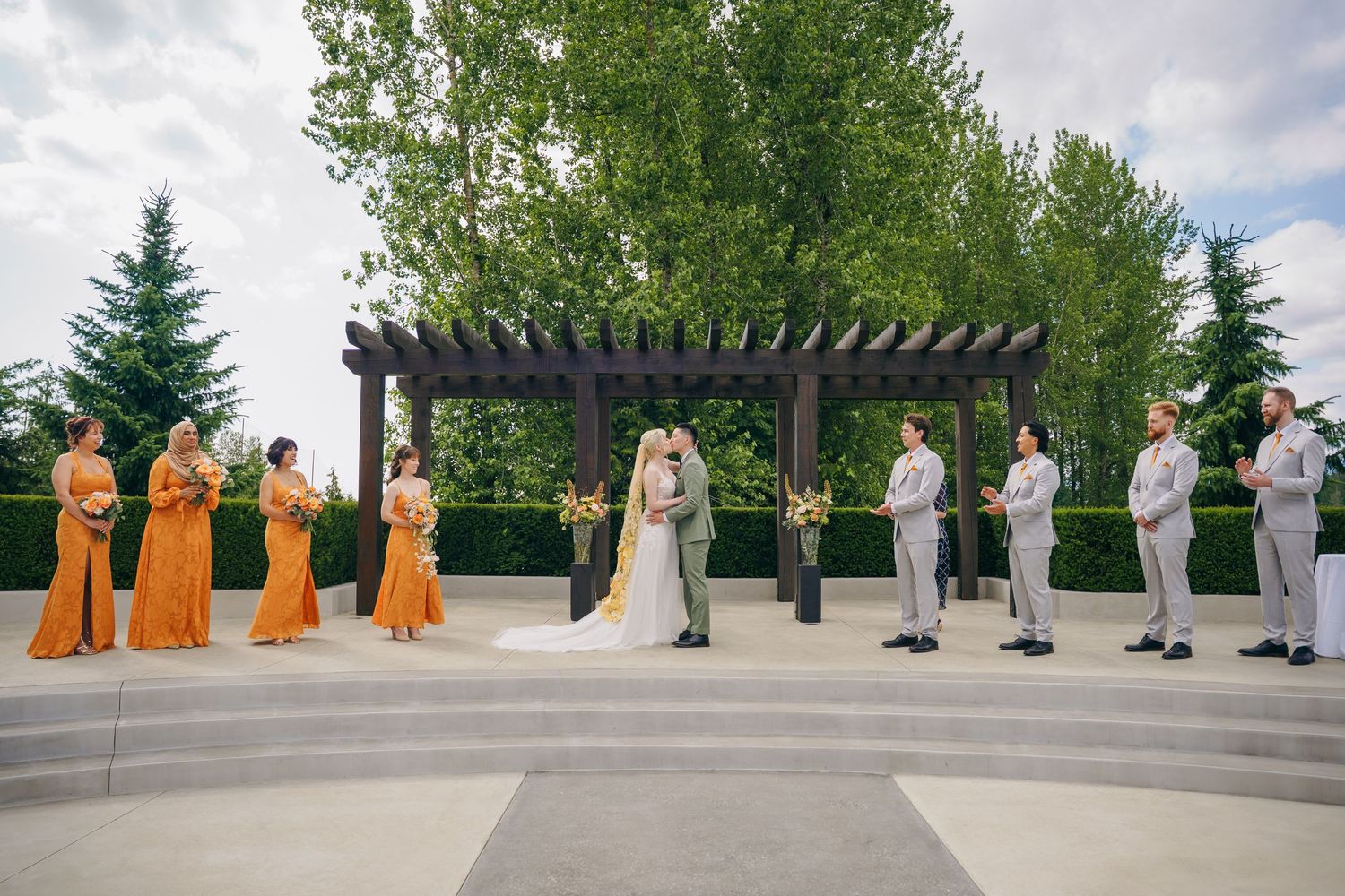 Wedding party lineup with bridesmaids in orange dresses against an outdoor landscape.
