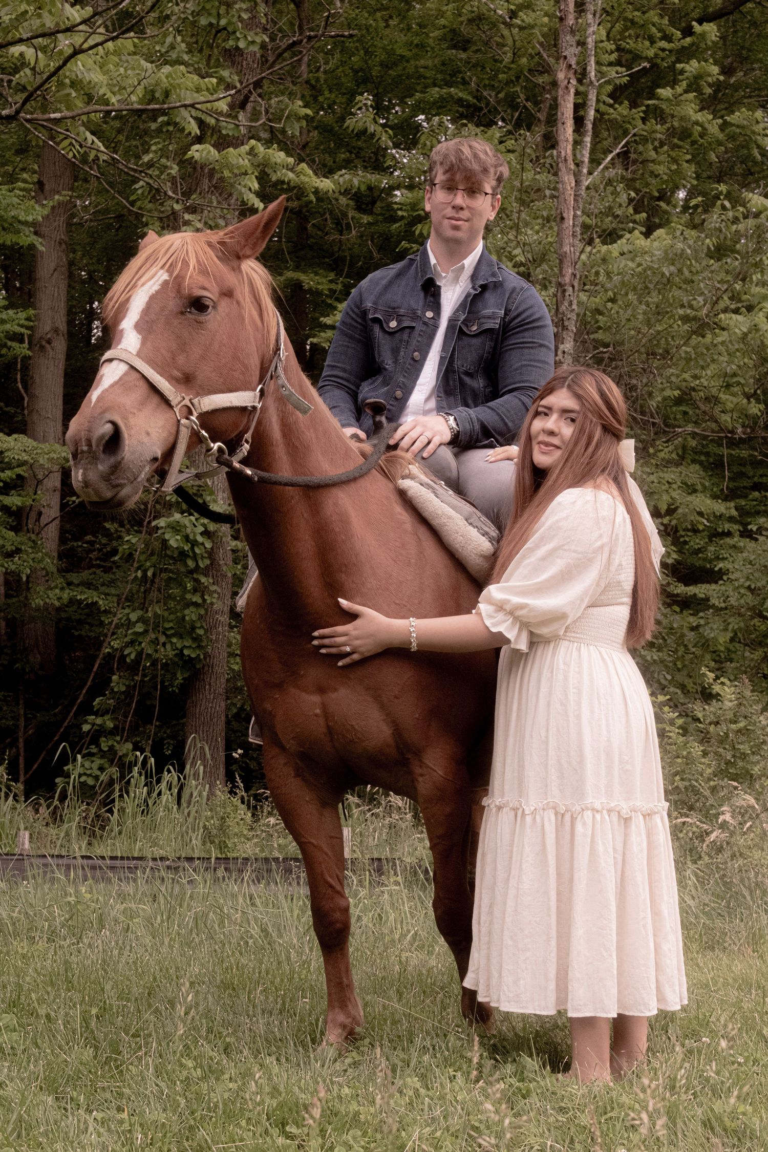 Couple poses with a brown horse in a rustic outdoor setting with trees.