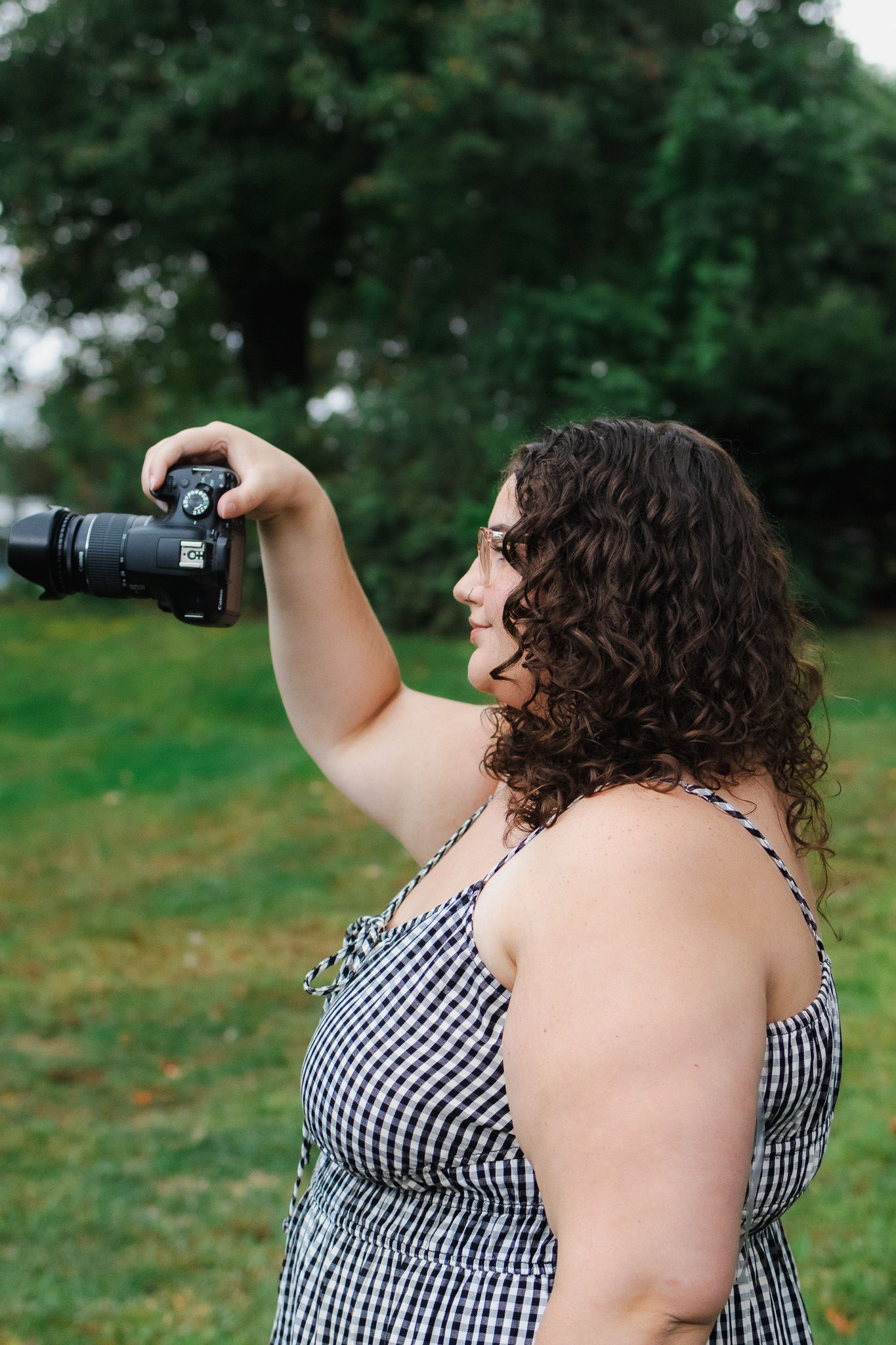 Person in gingham dress taking photos outdoors with camera in grassy field on sunny day.