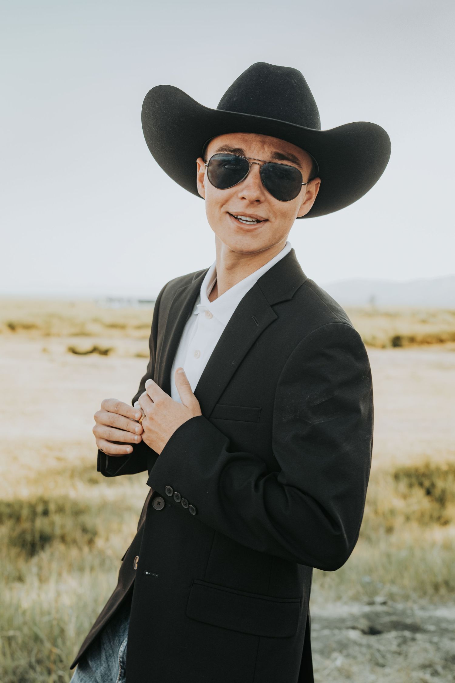 A person wearing aviator sunglasses, cowboy hat and black blazer poses in a rural field at sunset.