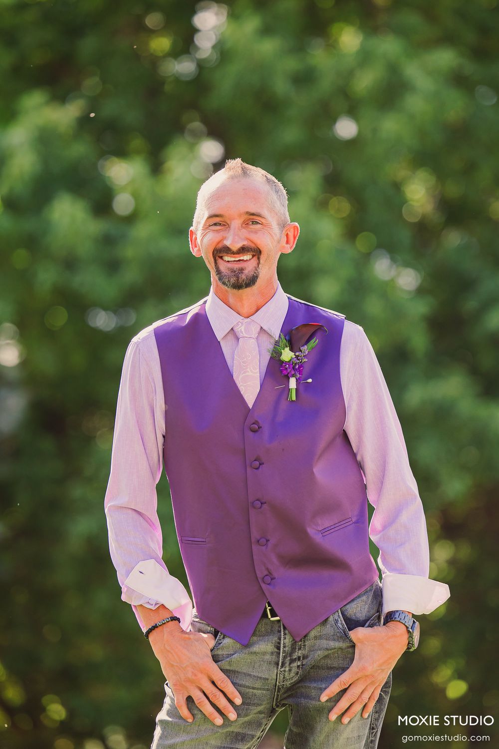 Wedding party member in purple vest and white shirt poses outdoors against green foliage background.