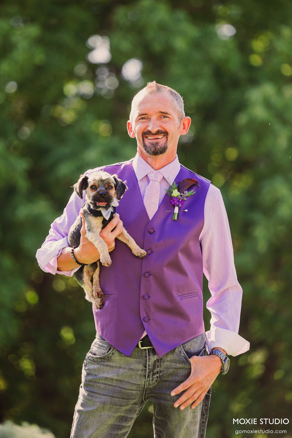 Groomsman in purple vest holds small puppies while smiling outdoors during wedding photoshoot.
