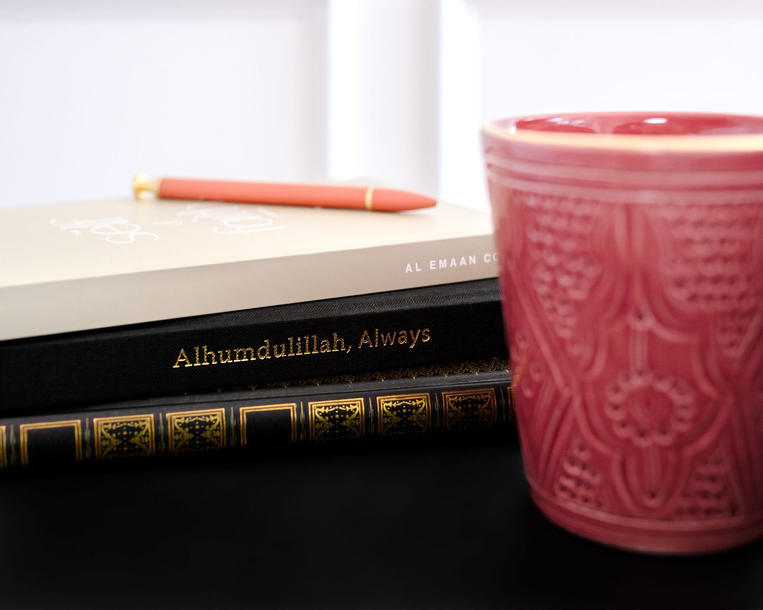 A hardcover book sits next to a pink ceramic mug and orange pencil on a dark surface.