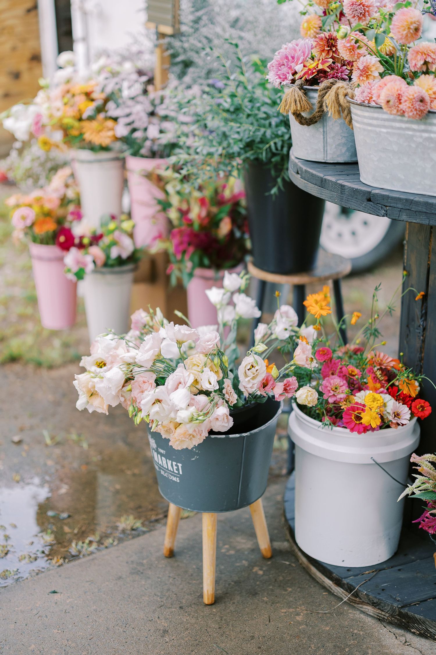 Colorful flower arrangements displayed in various pots and buckets on rustic shelves and stands at a garden shop.