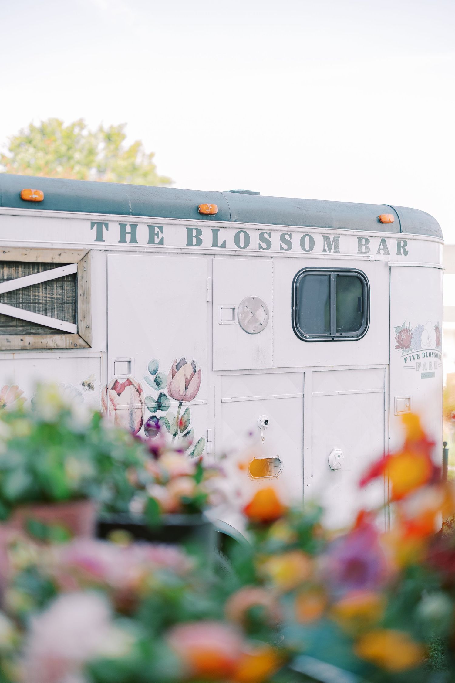 A vintage white trailer labeled 'The Blossom Bar' with colorful flowers in the foreground.