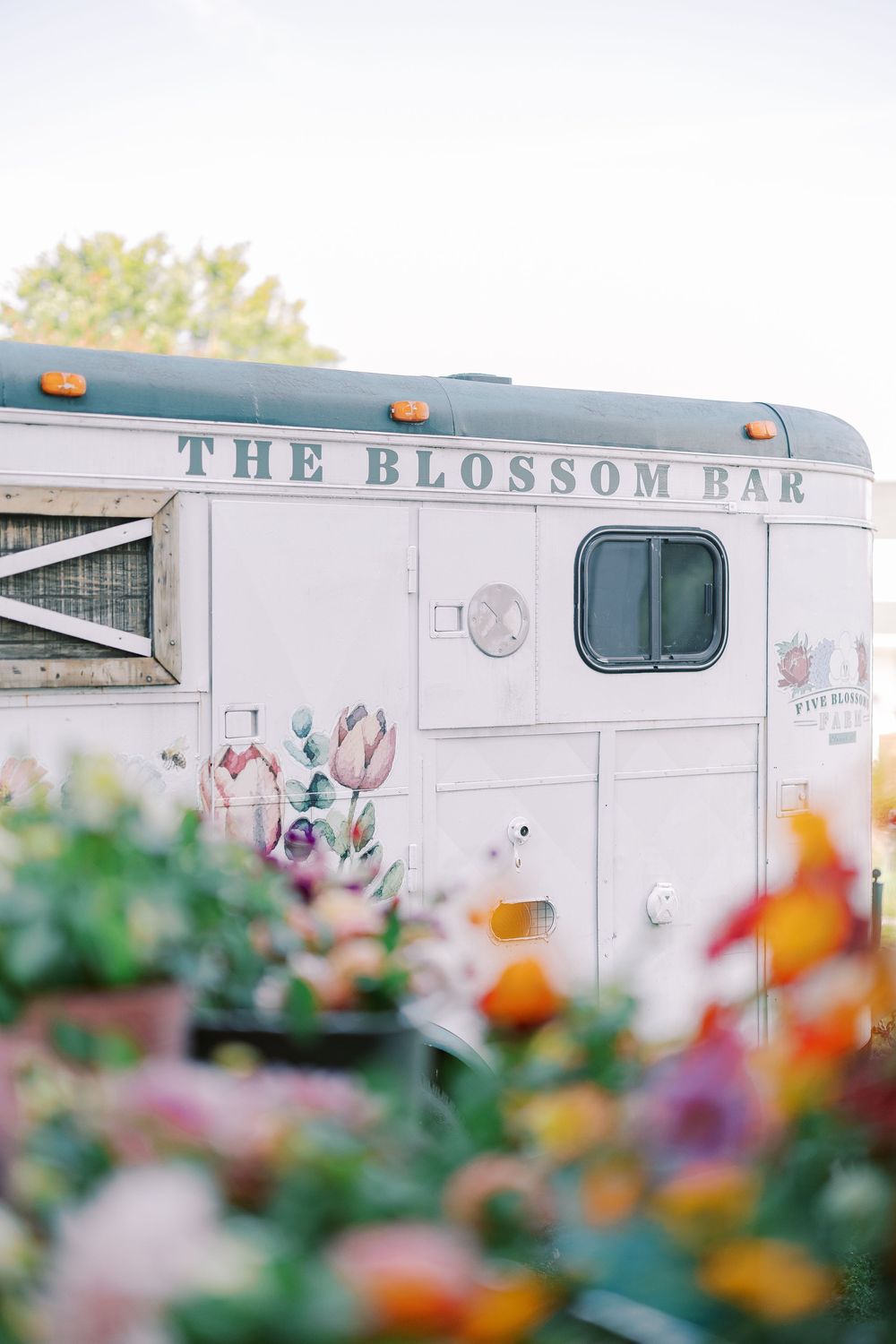 A vintage white trailer labeled 'The Blossom Bar' with colorful flowers in the foreground.