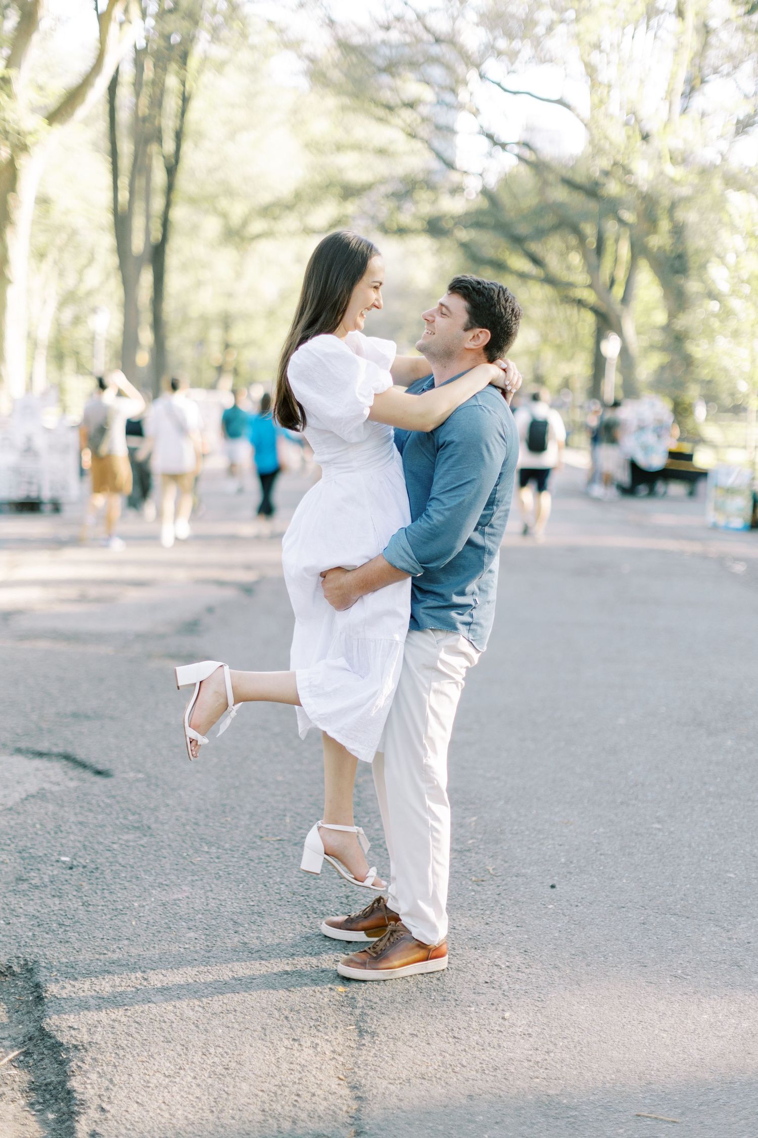 A couple in coordinated blue and white outfits embrace and dance together on a tree-lined pathway during a photoshoot.