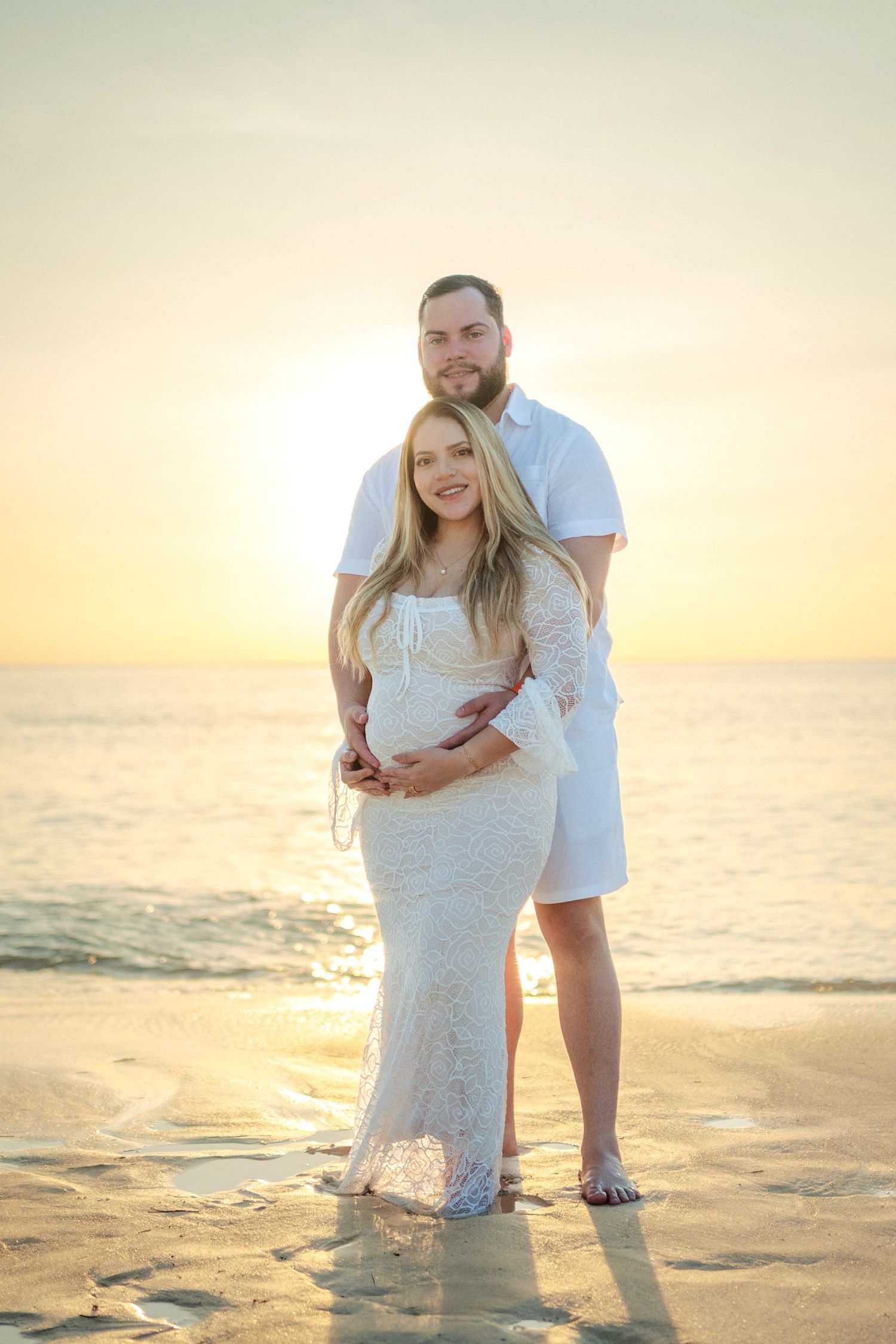 A couple embraces during sunset on the beach, with golden light reflecting off the ocean as waves lap at their feet.