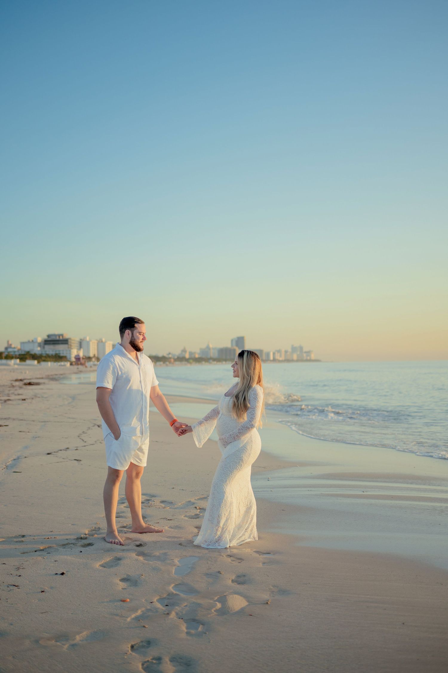 A couple in white outfits holds hands while walking along a scenic beach during sunset with a cityscape in the distance.