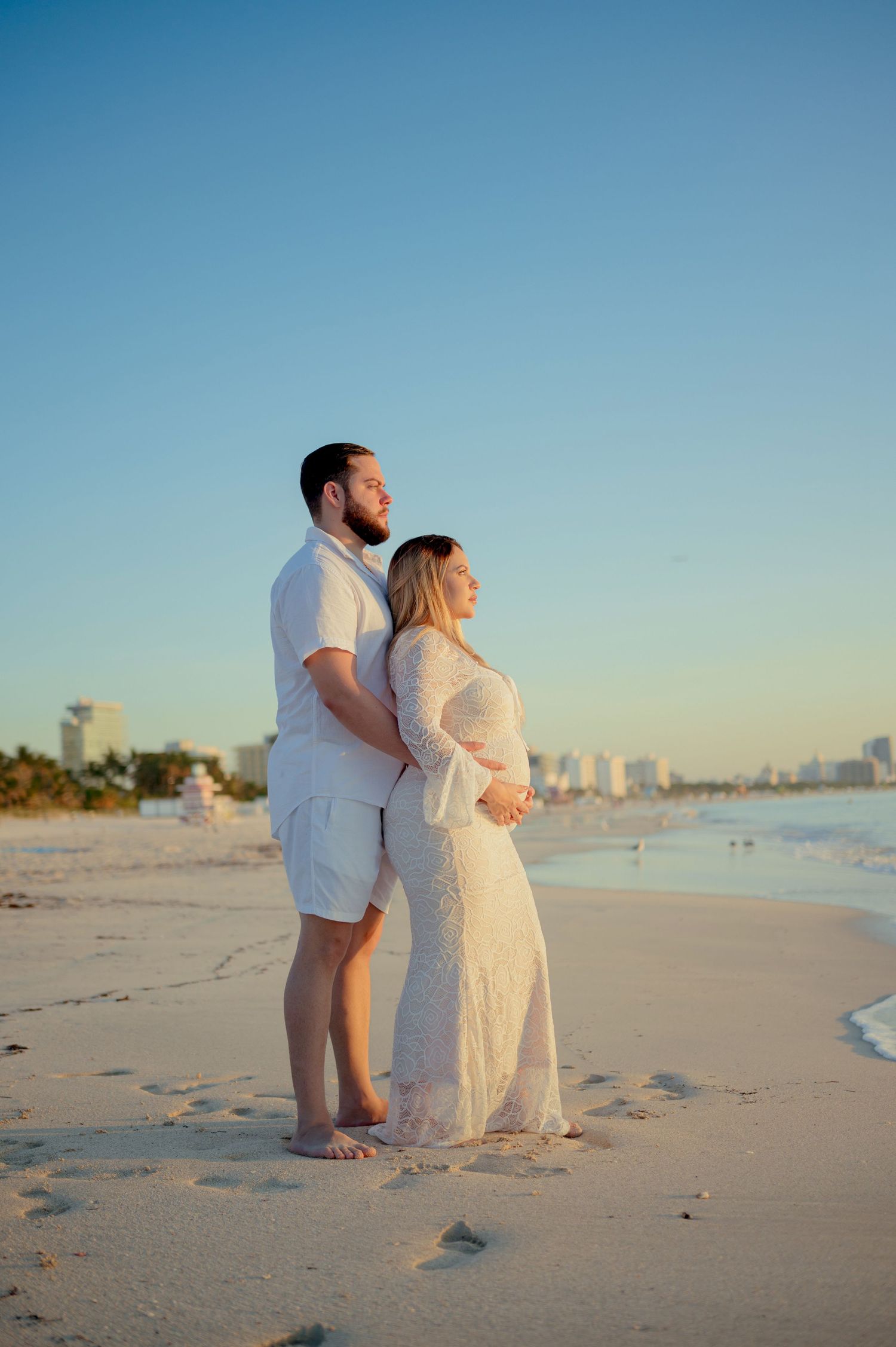 Couple in white attire standing on a sandy beach during sunset with cityscape in background.