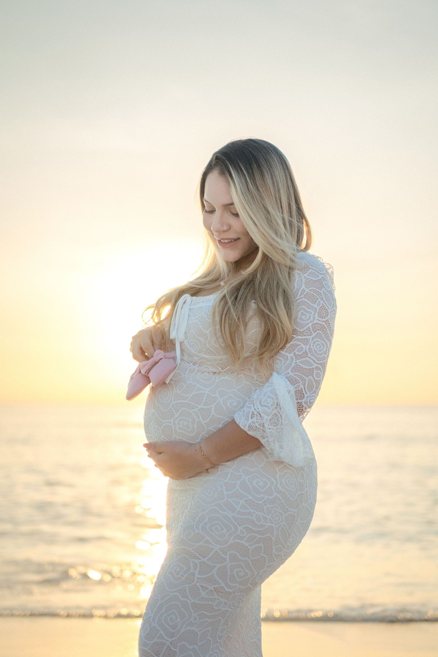 A pregnant woman in a white lace dress poses at the beach during golden hour sunset.
