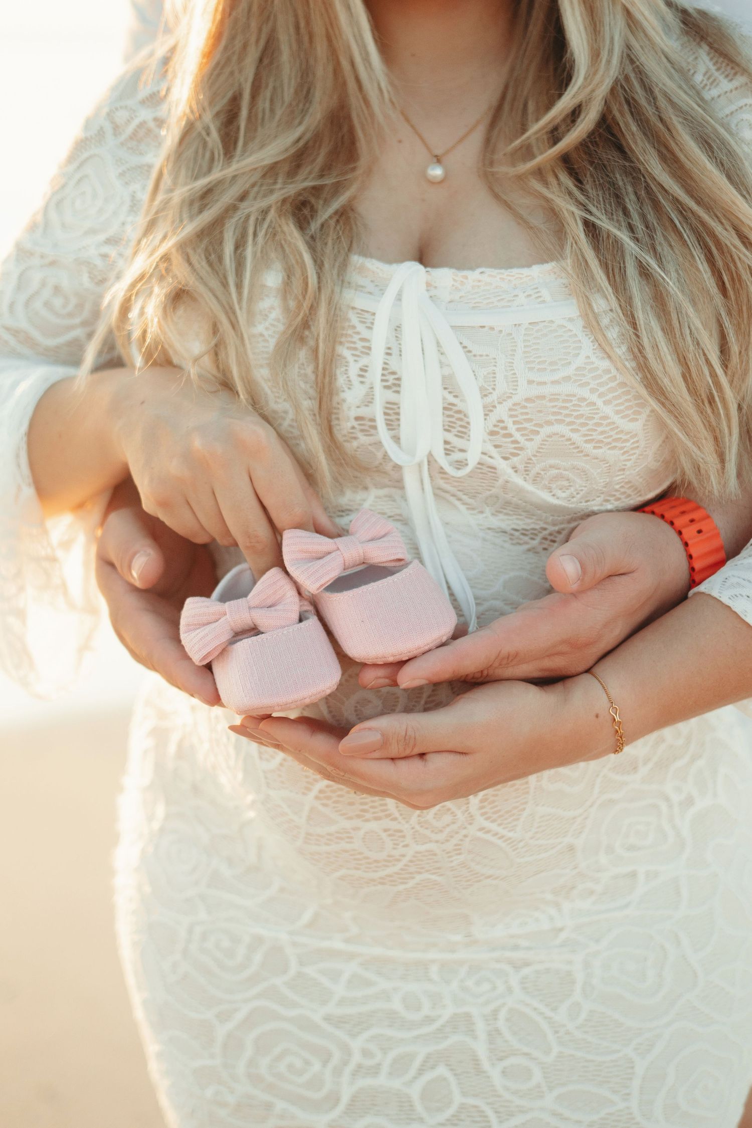 Person in white lace dress holds tiny pink baby shoes in their hands against sunlit backdrop.