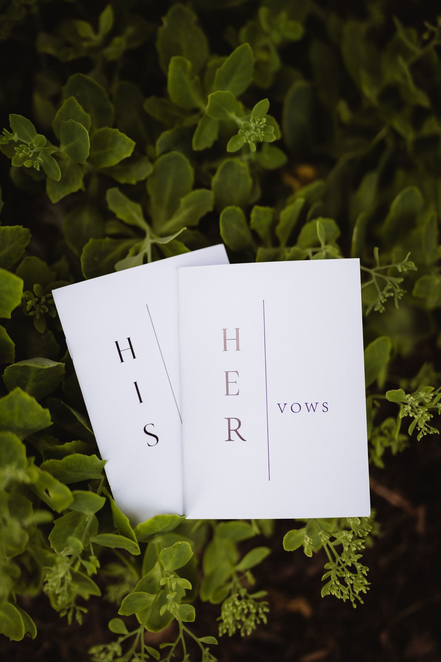 Three white wedding vow booklets with minimalist text 'HIS HER VOWS' placed on green foliage background.