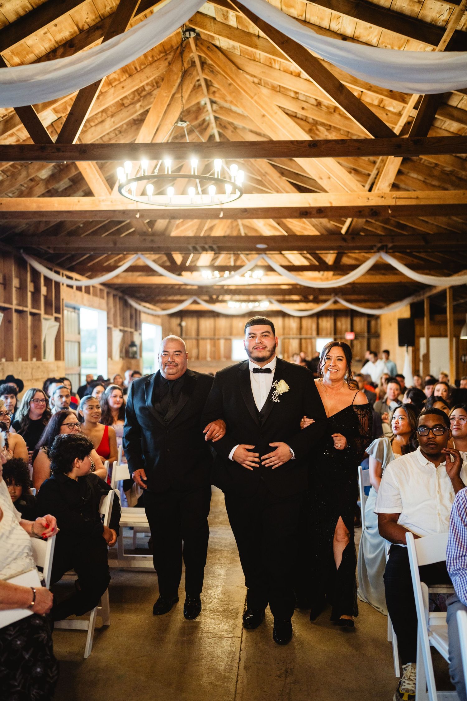 People walk down the aisle in a rustic wooden barn wedding venue decorated with white draping and chandeliers.