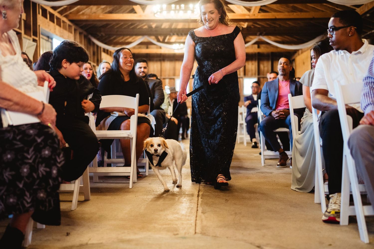 A dog walks down the aisle alongside someone in a formal black dress at an indoor wedding ceremony.