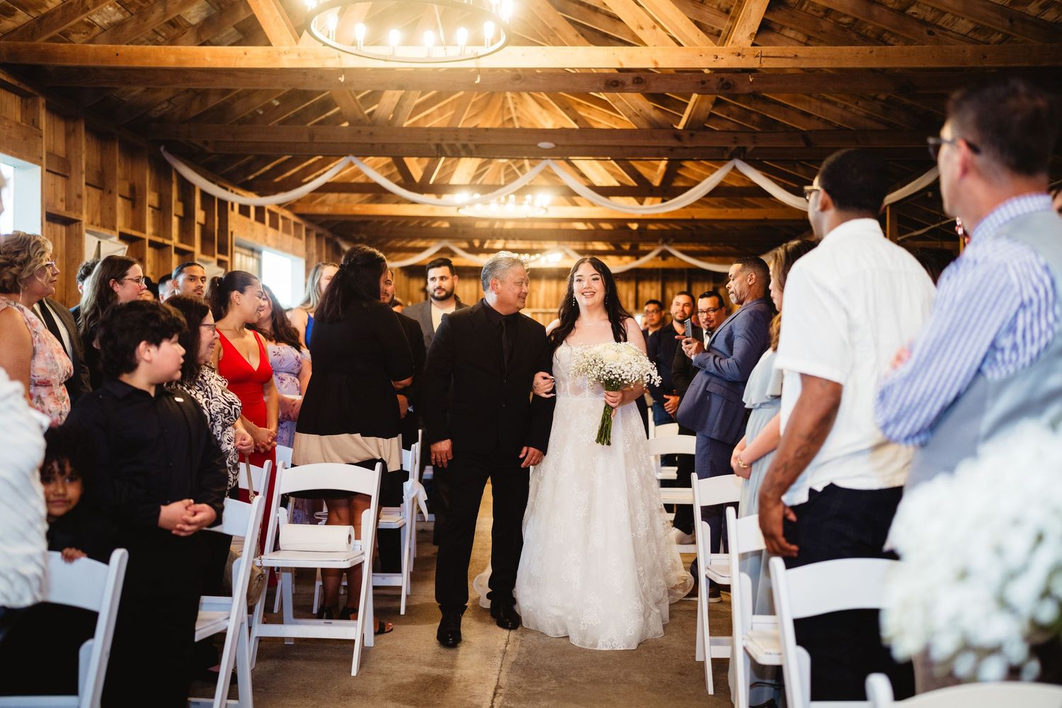 Wedding ceremony takes place in a rustic wooden barn with exposed beams and string lights overhead.