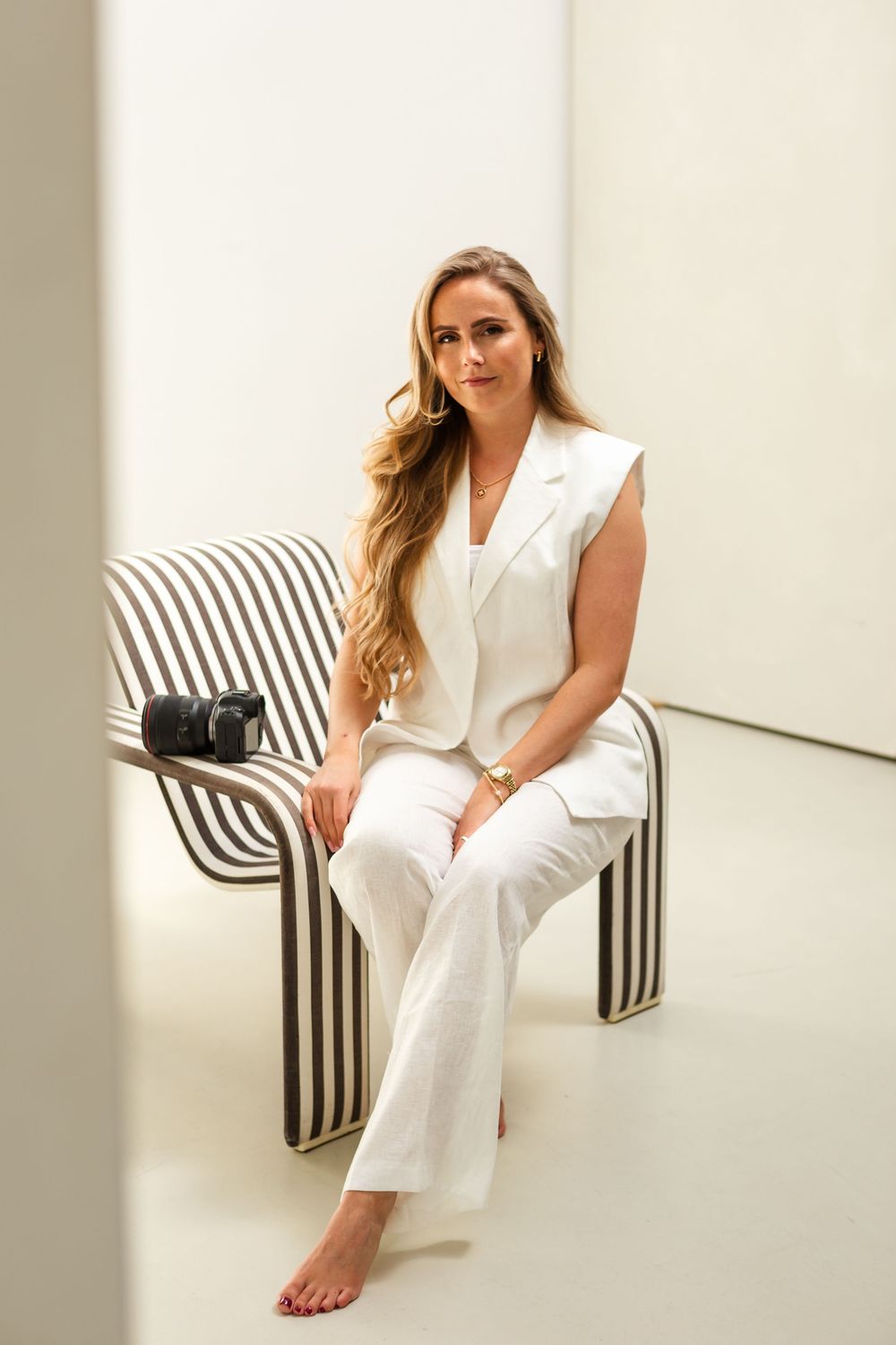 Professional model wearing all-white sleeveless suit ensemble sits elegantly on striped bench with black handbag.