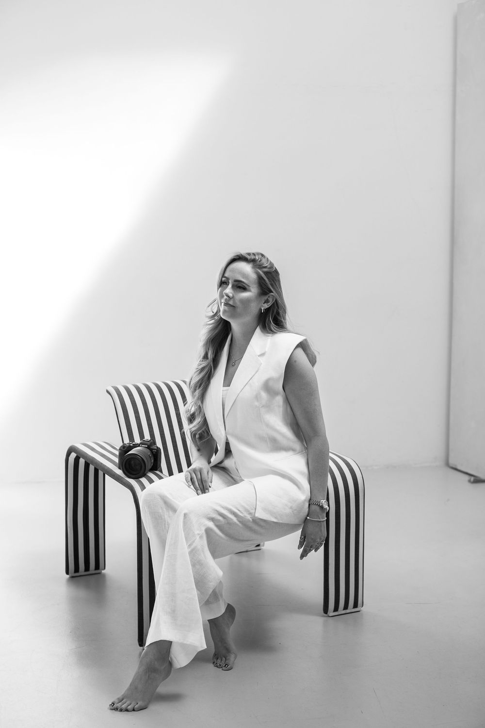A figure in a white pantsuit sits barefoot on a striped black and white bench against a minimalist white background.