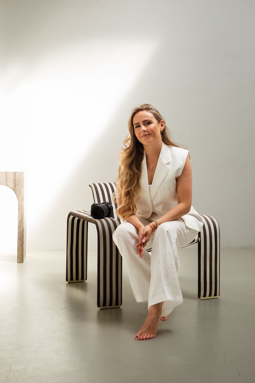 A person in an elegant white pantsuit sits barefoot on a striped modern chair in a minimalist studio setting.