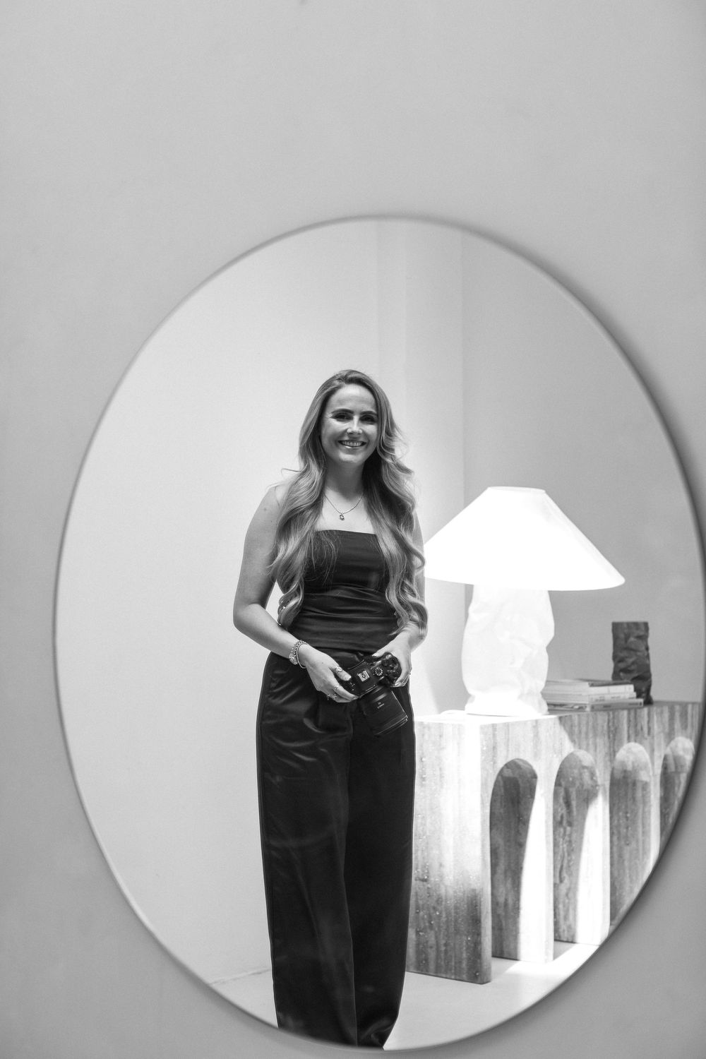 Black and white photograph of a figure in a formal evening gown standing beside a decorative table lamp in a mirror reflection.