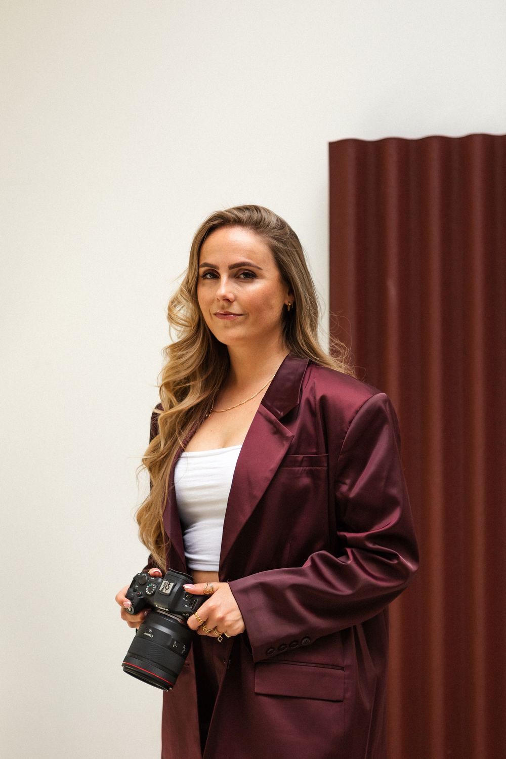 Professional photographer in burgundy blazer and white top holds DSLR camera against white wall and maroon curtain.