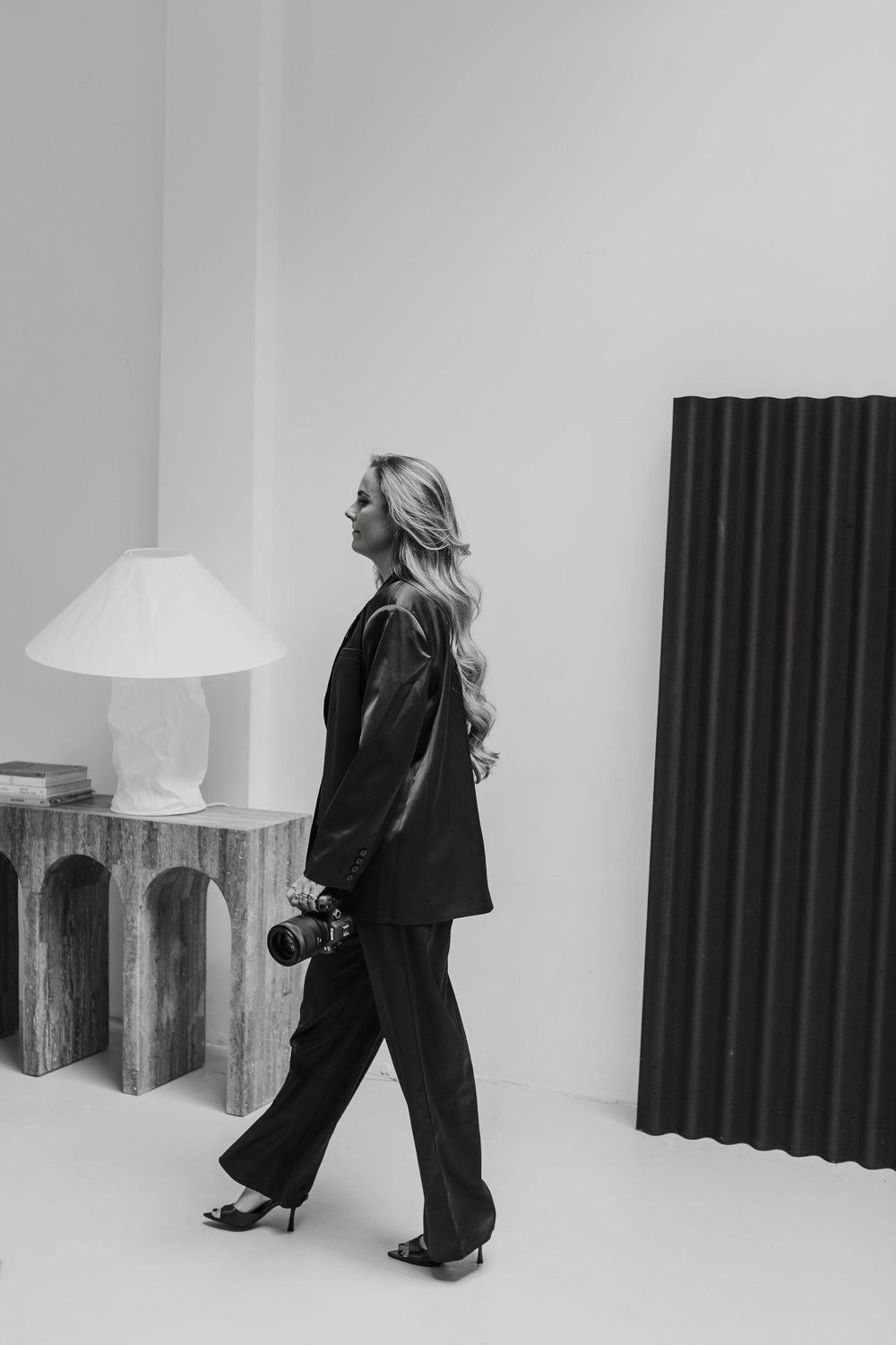 A figure in a black outfit and blonde hair walks in profile while holding a camera in a minimalist white studio setting.
