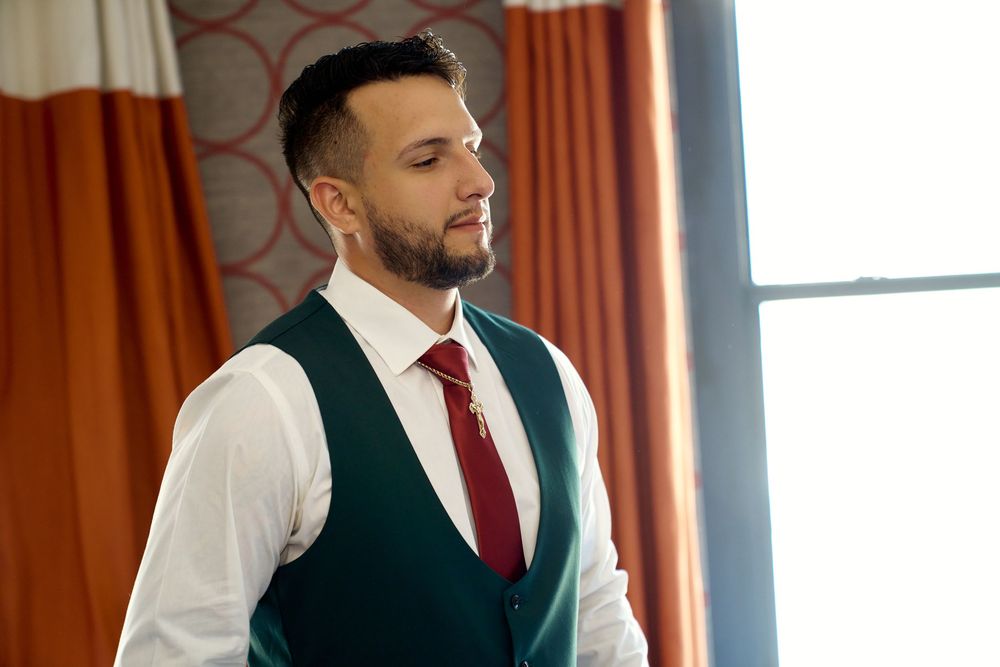 Professional in formal attire with vest and red tie looking contemplatively by window with orange curtains.