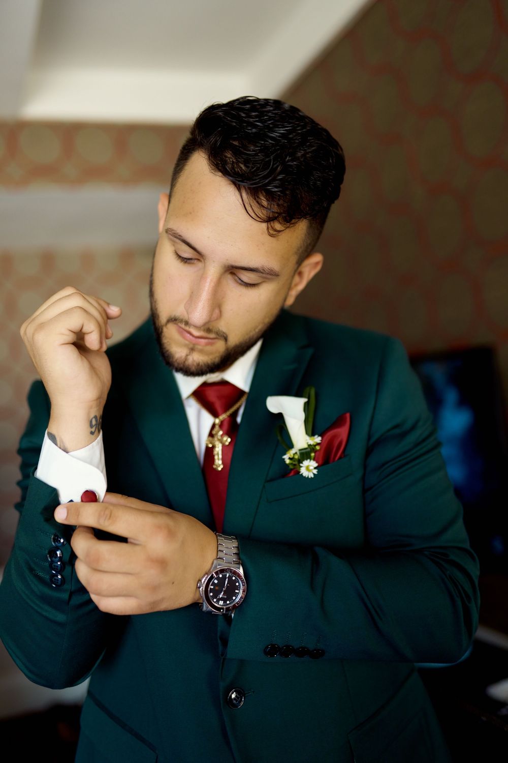 Groom in dark green suit with red tie and white boutonniere checks phone while getting ready for wedding ceremony.