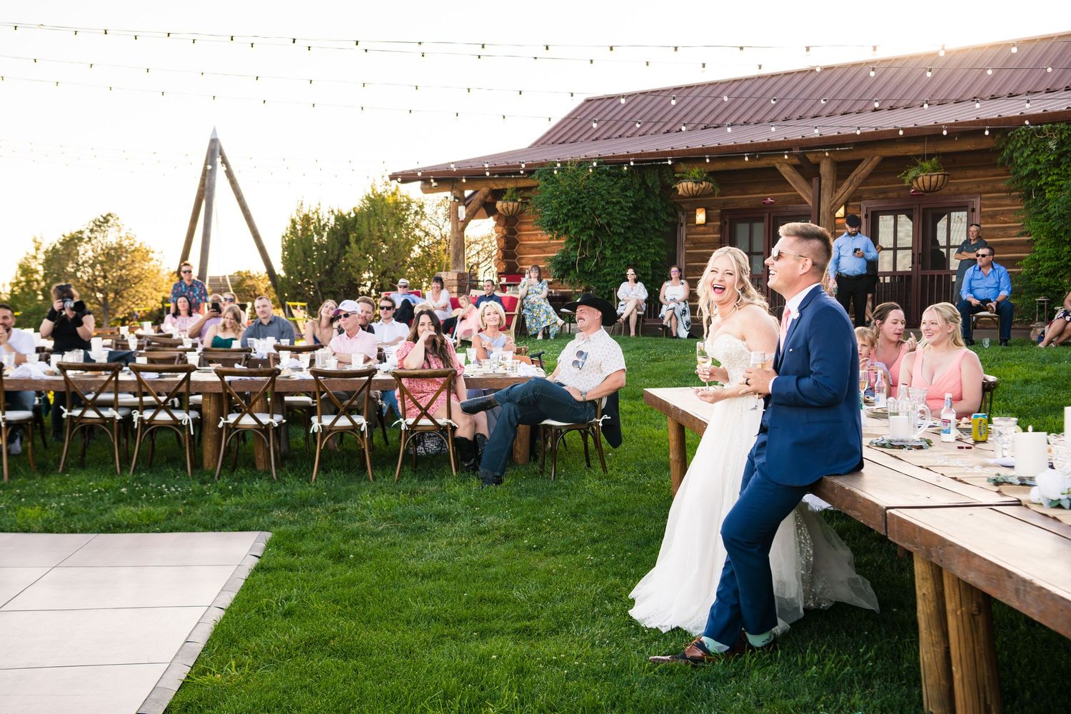 A panorama of wedding reception guests seated at long tables outdoors at a rustic venue during golden hour.
