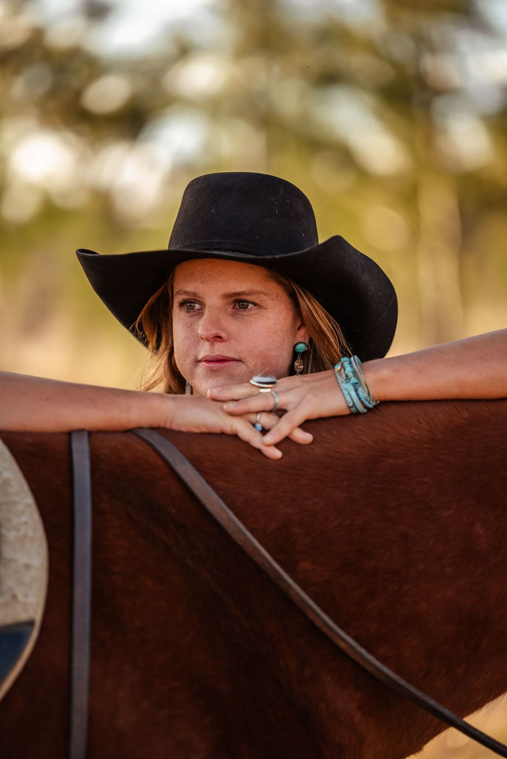 A figure in a black cowboy hat leans against a brown horse during a golden hour photoshoot.