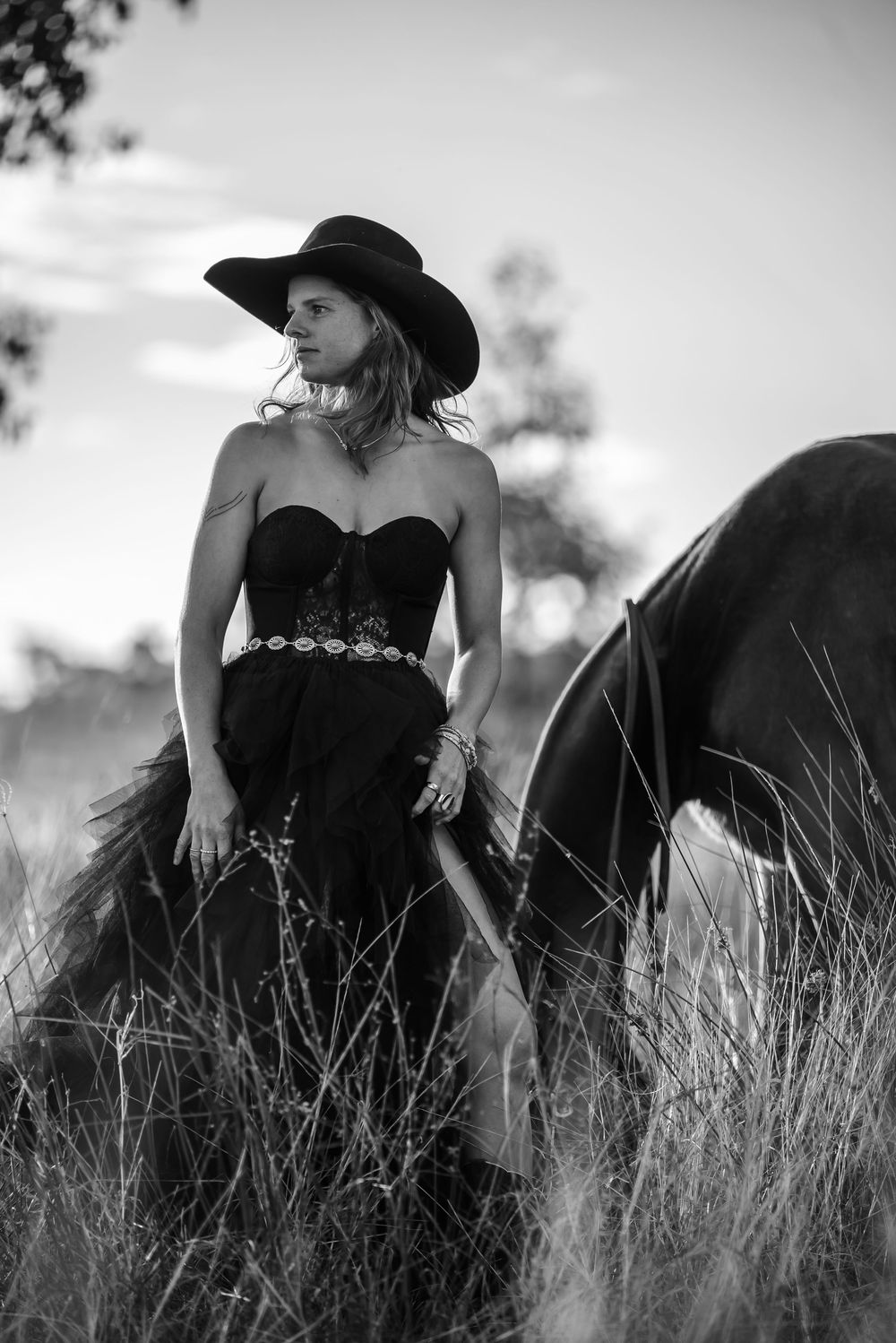 Artistic black and white photo of a figure in a black dress and hat among tall grass.