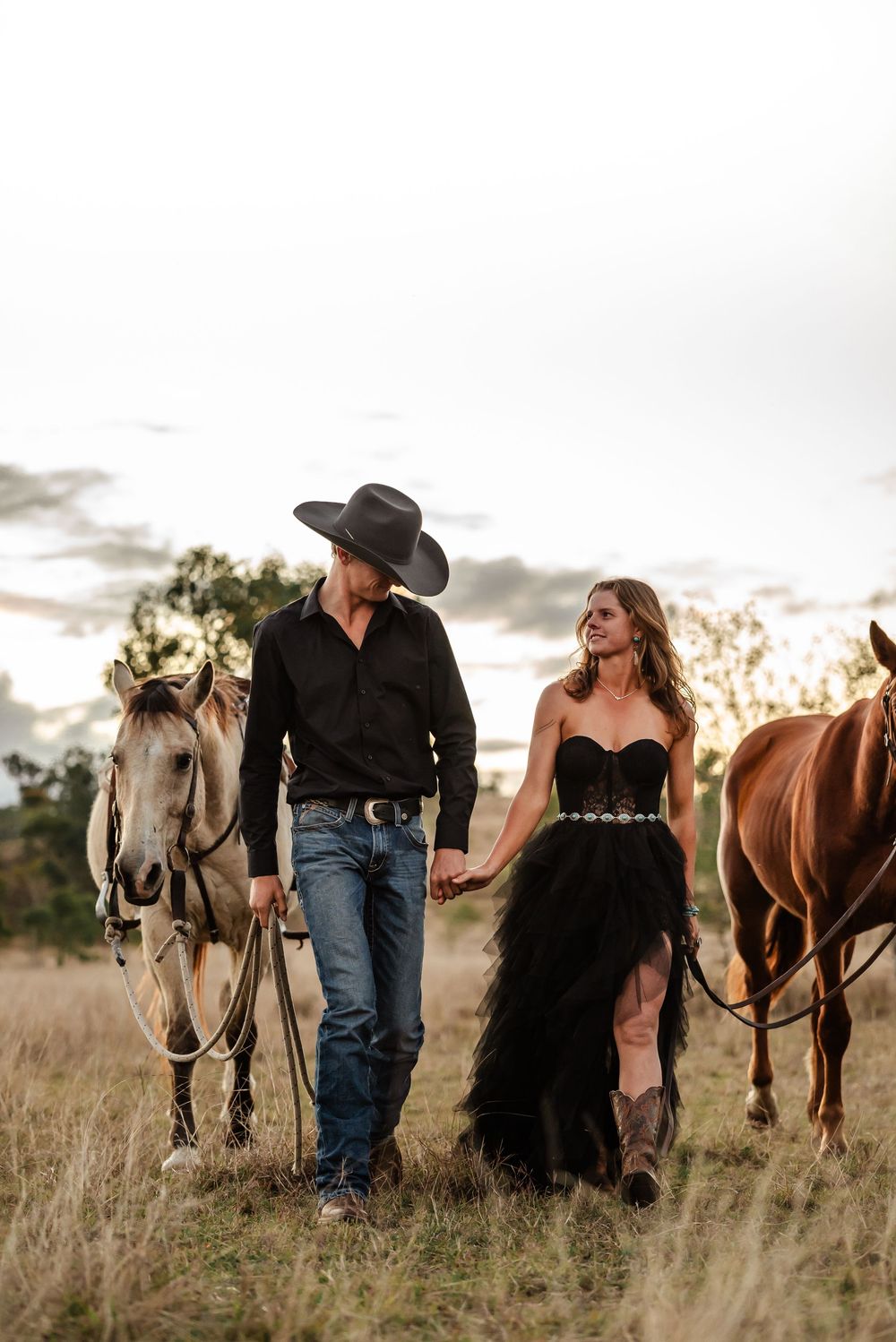 A couple walks with their horses through tall grass at sunset in western attire and formal dress.