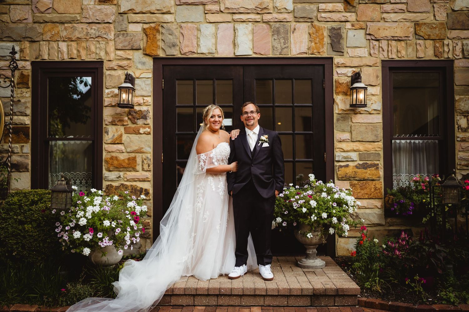 Wedding couple poses by rustic stone wall at elegant venue with dramatic lighting and floral arrangements.
